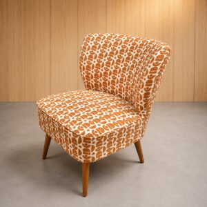 Poltroncina vintage con rivestimento in tessuto a fantasia geometrica arancione e bianca, gambe affusolate in legno chiaro.
