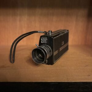 Cinepresa Super 8 vintage nera con obiettivo cromato e cinghia da polso, posizionata su una mensola di legno.