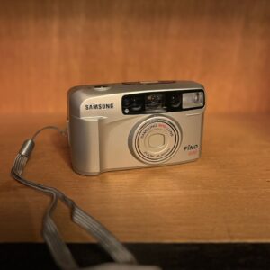 Fotocamera analogica compatta Samsung Fino 800 color argento con obiettivo zoom e flash, posizionata su una superficie di legno.