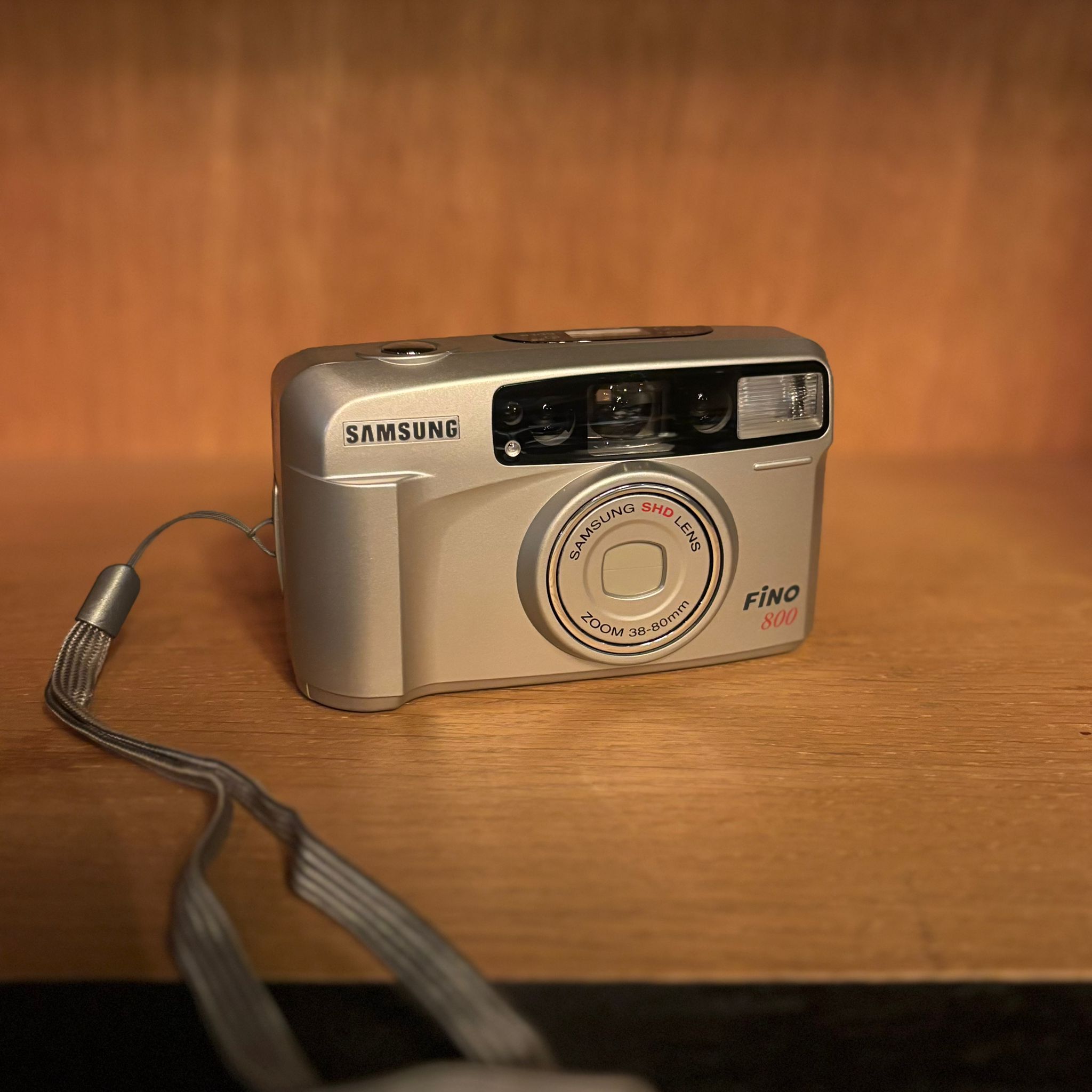 Fotocamera analogica compatta Samsung Fino 800 color argento con obiettivo zoom e flash, posizionata su una superficie di legno.