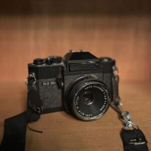 Fotocamera vintage nera SLX 500 con obiettivo Rikenon 50mm e tracolla, posizionata su una superficie di legno.