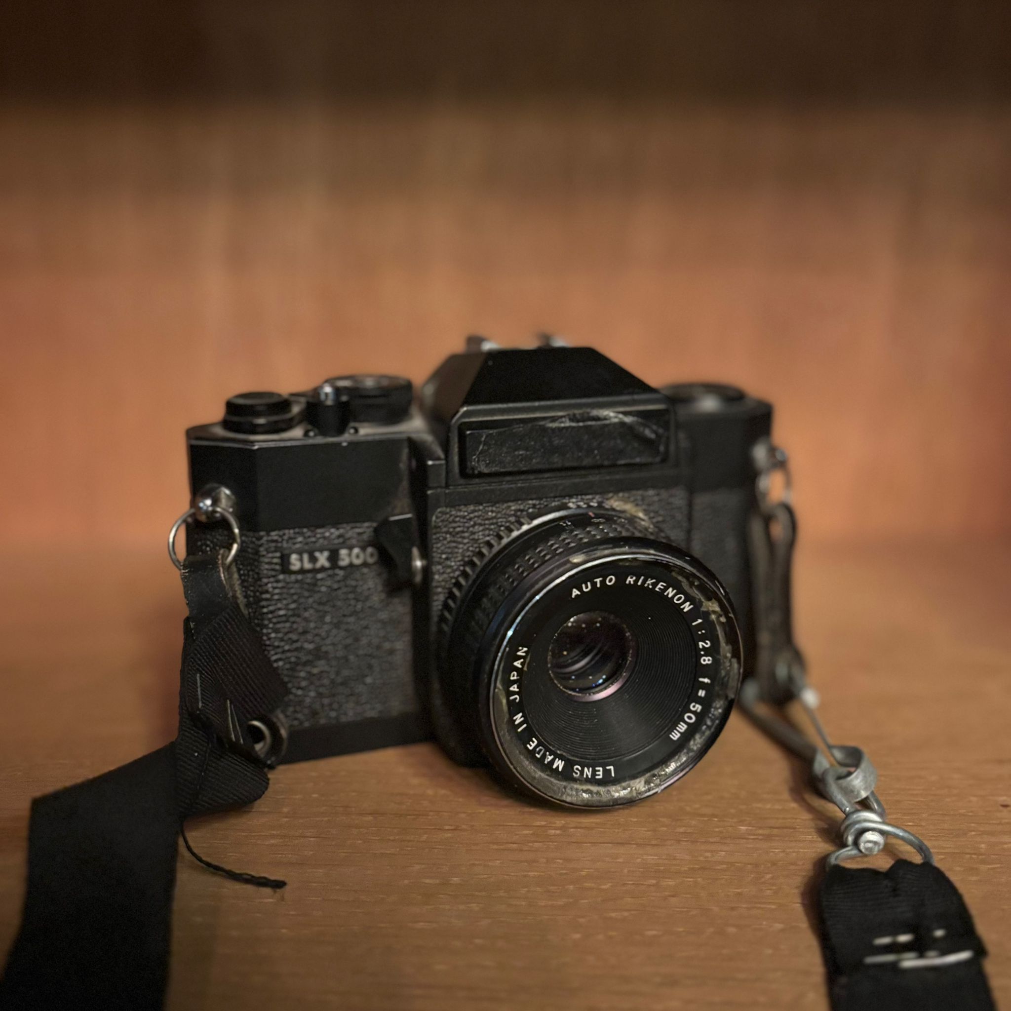 Fotocamera vintage nera SLX 500 con obiettivo Rikenon 50mm e tracolla, posizionata su una superficie di legno.