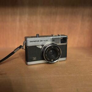 Fotocamera vintage Olympus-35 ECR con corpo cromato e rivestimento nero, obiettivo E. ZUIKO, su superficie di legno.