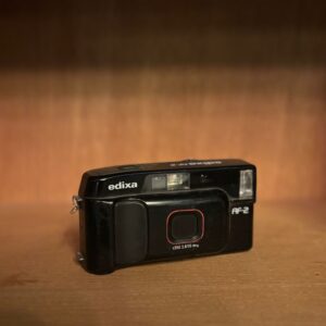 Fotocamera compatta Edixa AF-2 nera vintage con lente 3.8/35mm e flash integrato, posizionata su una superficie di legno.