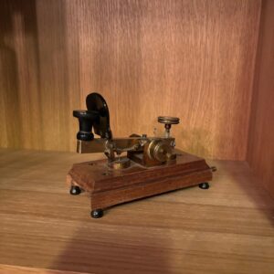 Manipolatore telegrafico vintage con base in legno scuro e meccanismi in ottone patinato, su uno scaffale di legno.