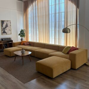 Ampio divano modulare giallo senape a L con pouf separato, posizionato in un soggiorno luminoso con finestra alta e tendaggi chiari.