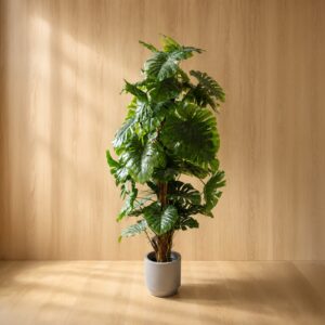 Grande pianta artificiale di Monstera con foglie verdi lussureggianti in un vaso cilindrico grigio effetto cemento, su sfondo di legno chiaro.
