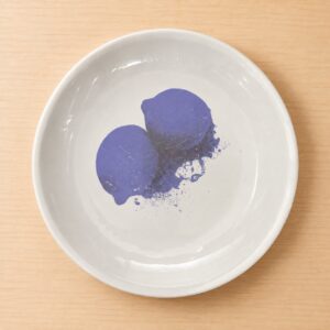 Piatto rotondo in ceramica bianca con bordo rialzato, decorato al centro con una stampa stilizzata e leggermente rovinata di due limoni viola e schizzi, su un tavolo di legno chiaro.