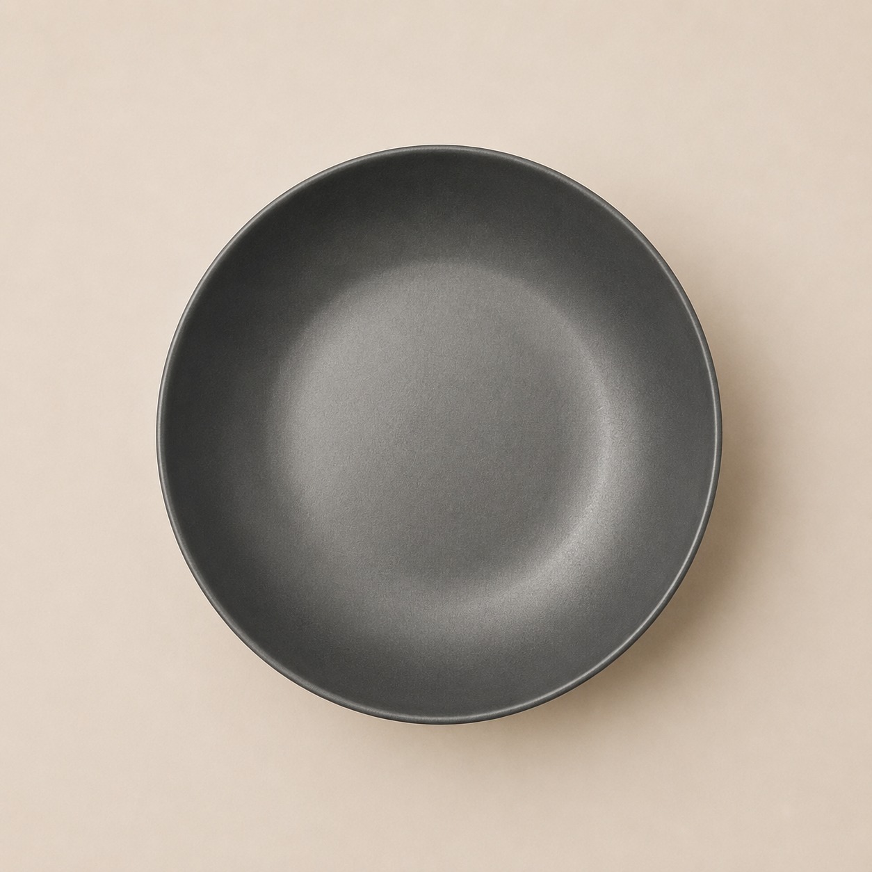 Piatto fondo rotondo di colore grigio antracite con finitura opaca, visto dall'alto su sfondo beige chiaro.