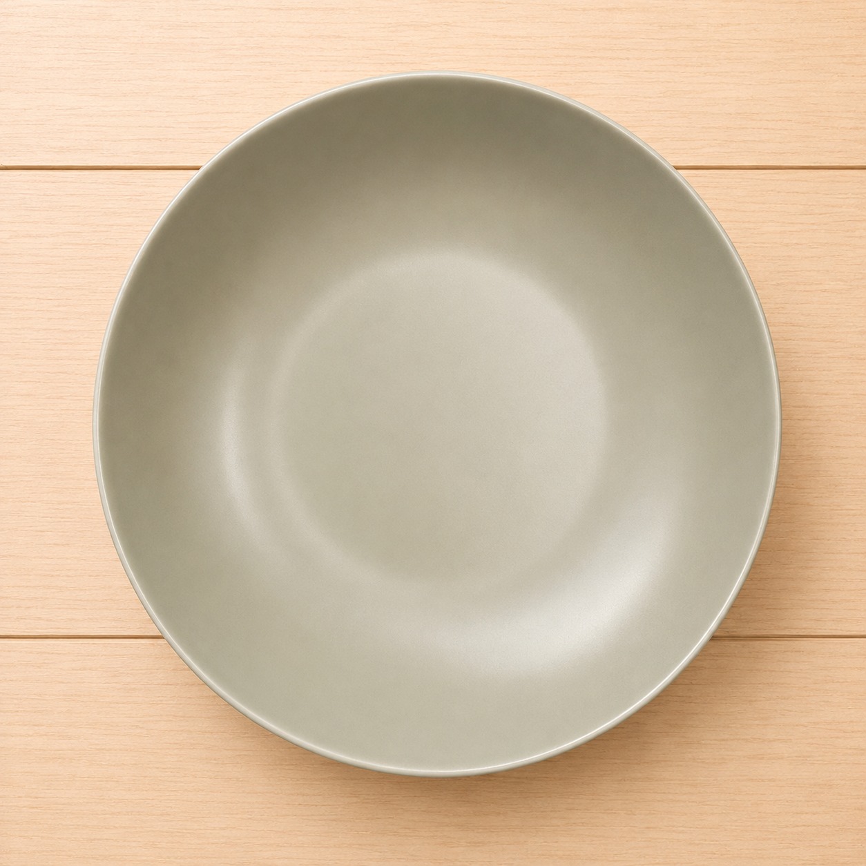 Piatto fondo rotondo di colore grigio-verde opaco, posizionato su un tavolo di legno chiaro con venature orizzontali.