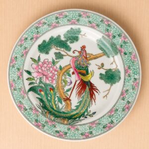 Piatto decorativo in ceramica bianca con pittura orientale di una fenice colorata, una peonia rosa e rami di pino.