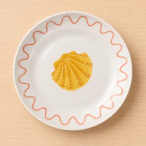Piatto in ceramica bianca con una conchiglia di capesante gialla al centro e un bordo ondulato rosa su un tavolo di legno chiaro.