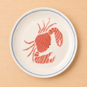 Piatto rotondo in ceramica bianca con bordo blu e illustrazione centrale di un granchio rosso stilizzato, su tavolo in legno chiaro.