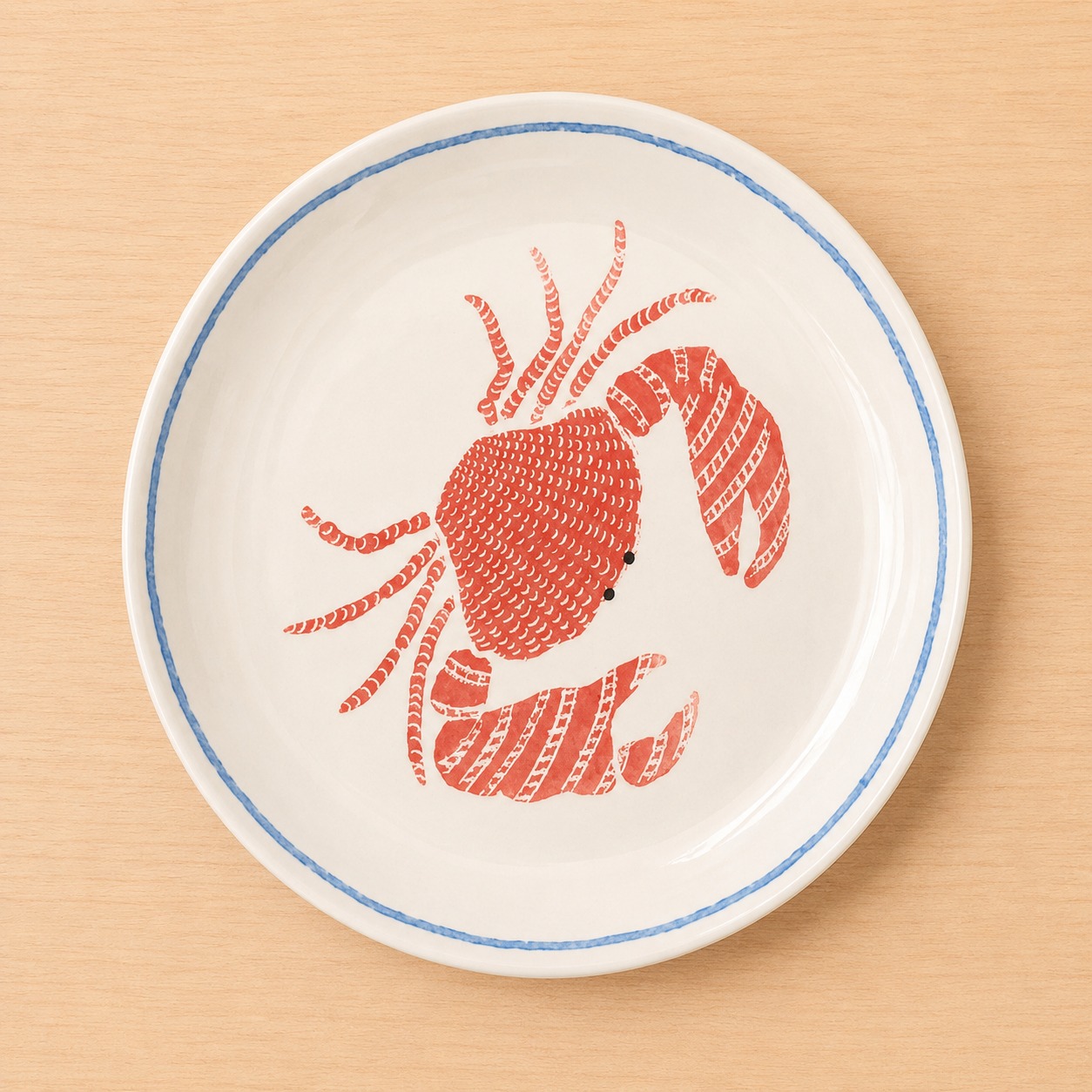 Piatto rotondo in ceramica bianca con bordo blu e illustrazione centrale di un granchio rosso stilizzato, su tavolo in legno chiaro.