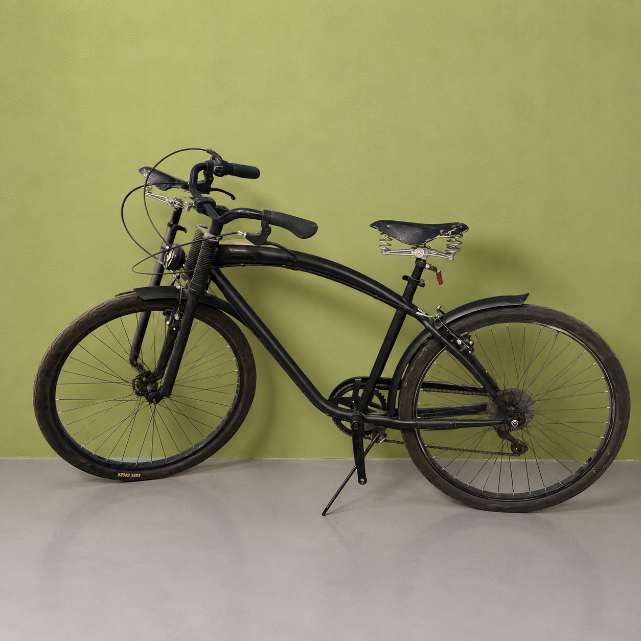 Bicicletta cruiser nera con telaio curvo, manubrio largo e sella a molle, appoggiata a una parete verde chiaro.