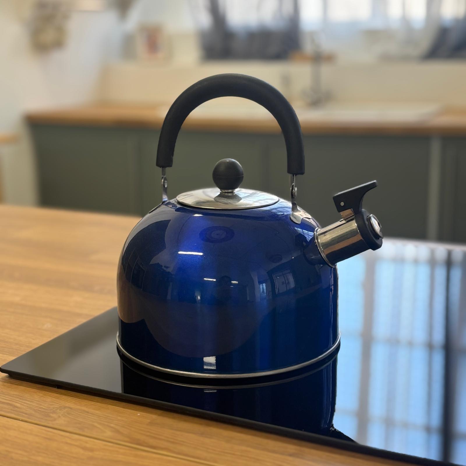Bollitore a fischio blu metallizzato con manico nero e dettagli cromati, posizionato su un piano cottura nero in una cucina moderna.