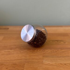 Barattolo di vetro sferico trasparente pieno di chicchi di caffè scuri, con coperchio metallico spazzolato, adagiato su un tavolo di legno.