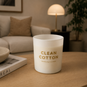 Candela decorativa bianca in vetro smerigliato con etichetta dorata "Clean Cotton" su un tavolino di legno in un ambiente domestico.
