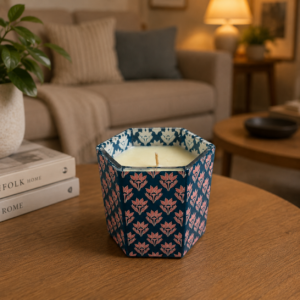 Candela decorativa in contenitore esagonale blu scuro con motivo floreale rosa, posta su un tavolino di legno in un ambiente domestico.