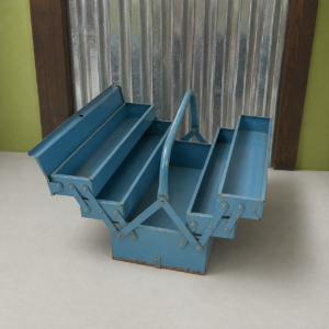 Cassetta degli attrezzi vintage in metallo blu aperta, con scomparti a fisarmonica e segni di usura, su sfondo industriale.