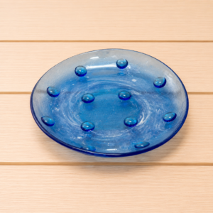 Piatto decorativo ovale in vetro blu intenso con rilievi a bolle sulla superficie, posizionato su un tavolo di legno chiaro.