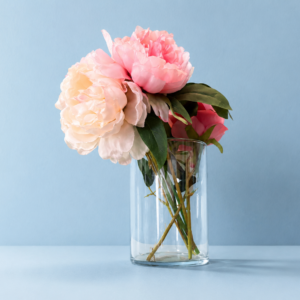Bouquet di peonie artificiali rosa e crema in un vaso di vetro trasparente su sfondo azzurro chiaro.