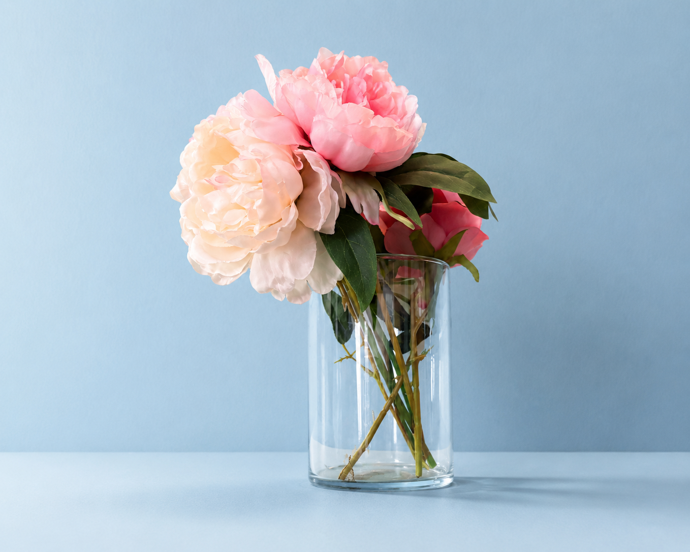 Bouquet di peonie artificiali rosa e crema in un vaso di vetro trasparente su sfondo azzurro chiaro.