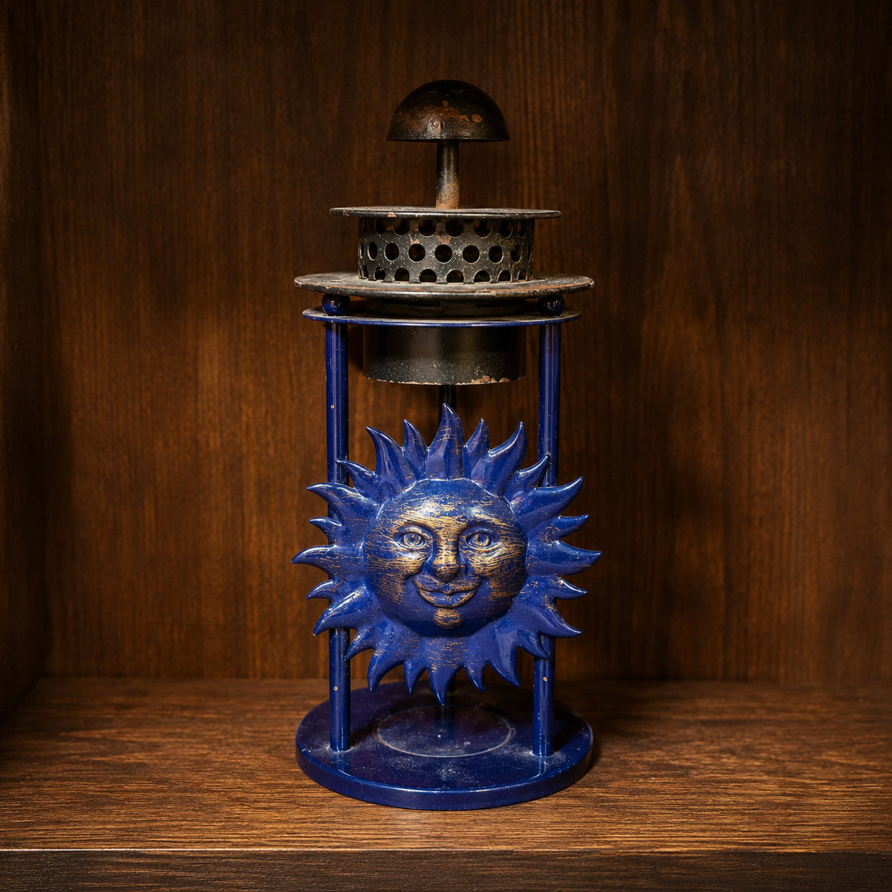 Bruciatore decorativo in metallo blu con un volto solare dorato, parte superiore patinata e perforata, su uno scaffale di legno.