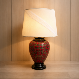 Lampada da tavolo con base a vaso rossa e motivo tartan, paralume crema con dettagli a pieghe, su sfondo in legno chiaro.