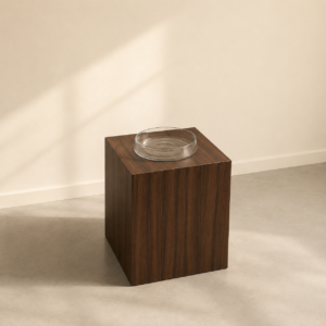 Piedistallo cubico in legno scuro con venature verticali, sormontato da un piatto in vetro trasparente, su pavimento chiaro con ombre.