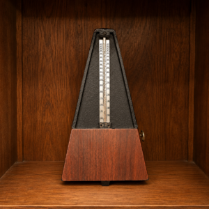 Metronomo classico con base in legno scuro e corpo piramidale nero, con asta graduata e peso, posizionato su una mensola di legno.