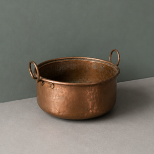 Calderone in rame antico con patina e due manici ad anello, posizionato su una superficie grigia davanti a una parete verde scuro.