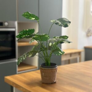 Pianta artificiale tropicale con foglie verdi variegate in un vaso rivestito di juta, posizionata su un piano di lavoro in legno.