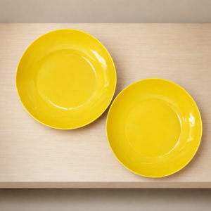 Due piatti fondi di colore giallo brillante, lucidi, posizionati su una mensola di legno chiaro, visti dall'alto.