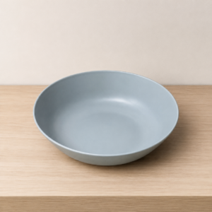 Ciotola piatta in ceramica di colore grigio azzurro opaco, posizionata su un tavolo di legno chiaro con sfondo beige.