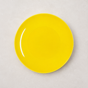 Piatto piano rotondo di colore giallo brillante con finitura lucida, posizionato centralmente su una superficie grigio chiaro.