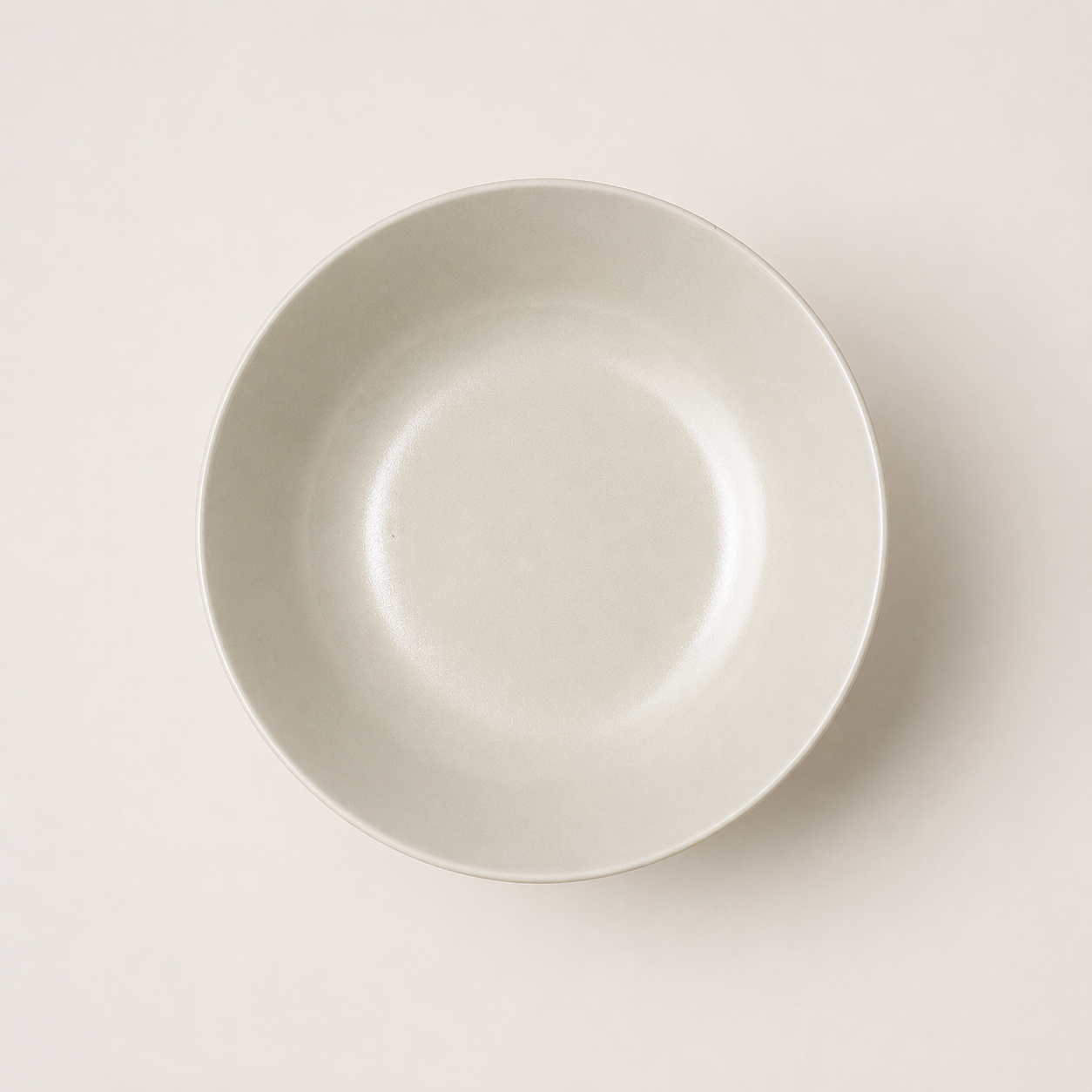 Ciotola piatta in ceramica o gres di colore beige chiaro, vista dall'alto, su sfondo neutro, con texture leggermente granulosa.