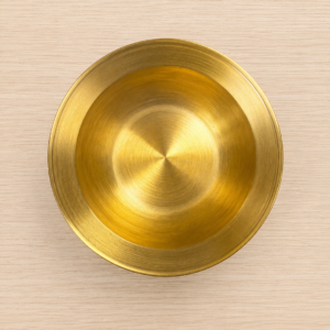 Piatto decorativo circolare in ottone dorato con finitura spazzolata, visto dall'alto su sfondo in legno chiaro.