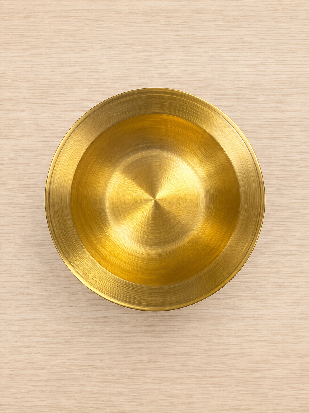 Piatto decorativo circolare in ottone dorato con finitura spazzolata, visto dall'alto su sfondo in legno chiaro.