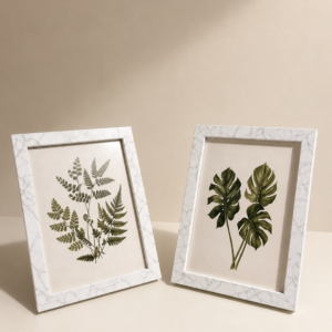 Due cornici bianche effetto marmo con stampe botaniche (felce e monstera) appoggiate su una superficie chiara.