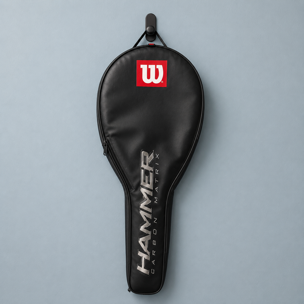 Fodero nero per racchetta da tennis Wilson Hammer Carbon Matrix appeso a un gancio nero su parete azzurra.