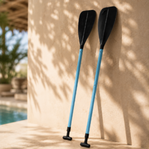 Due pagaie con aste blu e pale nere appoggiate a una parete chiara, con piscina e vegetazione sfocate sullo sfondo.