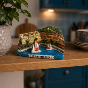 Miniatura tridimensionale colorata di Sanremo con edifici, colline, mare e barche a vela, su un ripiano di legno.