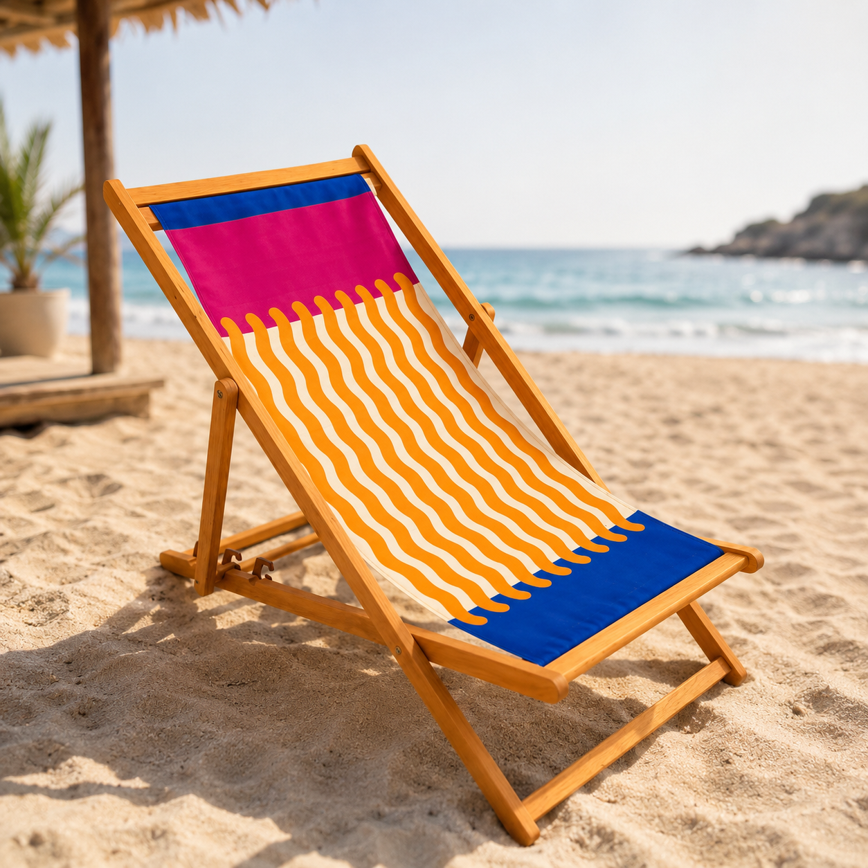 Sdraio pieghevole in legno chiaro con tessuto a fantasia geometrica fucsia, arancione a onde e blu, su una spiaggia sabbiosa.