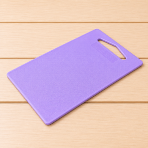 Tagliere in plastica di colore viola con superficie testurizzata e manico integrato, posizionato su un tavolo di legno chiaro.