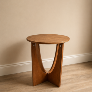 Tavolino laterale rotondo in legno chiaro con base a due elementi curvi, su pavimento in parquet e parete beige.