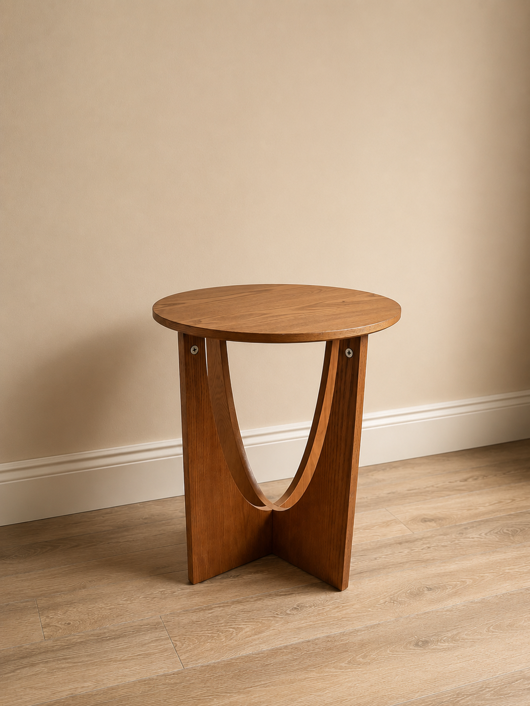 Tavolino laterale rotondo in legno chiaro con base a due elementi curvi, su pavimento in parquet e parete beige.