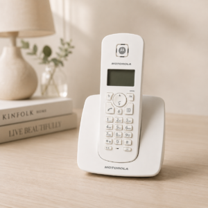 Telefono cordless Motorola bianco con base di ricarica su un tavolo di legno chiaro, con libri e lampada sfocati sullo sfondo.